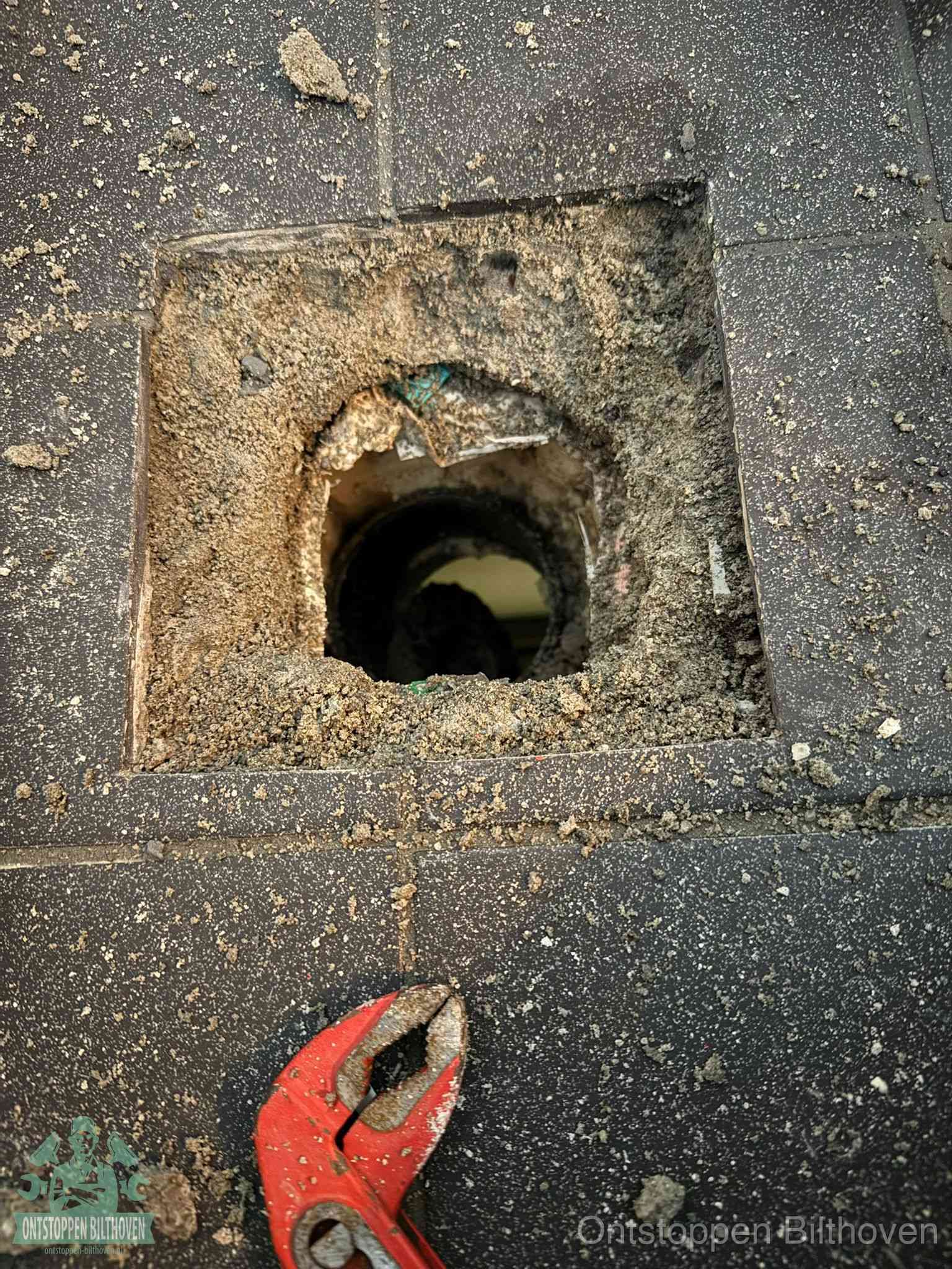 Een geopend rioolputje in een betontegelvloer met losgemaakt rooster en een zichtbare ronde afvoerbuis, het achterhalen waar de afvoerverstopping zit in Bilthoven