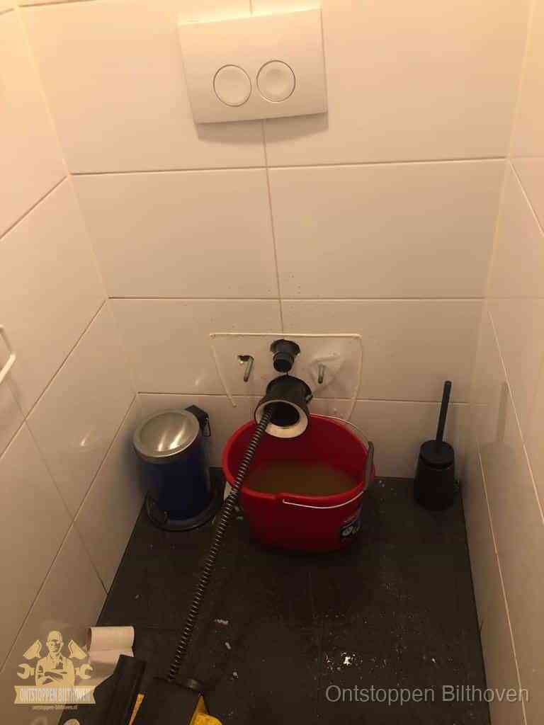 Een ontstopte wand-wc met verwijderde toiletpot waar een ontstoppingsveer in de wanduitgang verdwijnt, het achterhalen waar de verstopping zit in Bilthoven