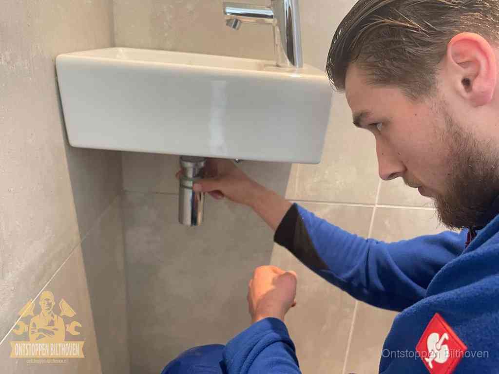 Een monteur draait met de hand de zwanenhalssifon onder een fonteinwasbak in de toiletruimte los om de bron van rioollucht in Bilthoven te vinden