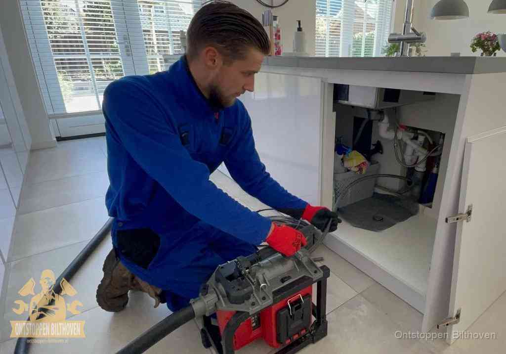 Een monteur voert een professionele elektrische ontstoppingsmachine in de afvoer onder de spoelbak tijdens het gootsteen ontstoppen in Bilthoven